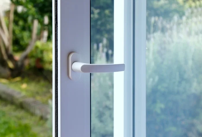 Close-up of a white window handle attached to a glass door, partially open. The background shows a blurred view of a garden with green foliage and a stone path, suggesting an outdoor setting on a bright day, enhanced by the craftsmanship of a local company specializing in repair.