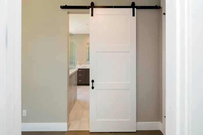 A white sliding barn door partially open reveals a modern bathroom in Tampa, FL, with a double vanity and large mirror. The room features neutral-colored walls and floor tiles, creating a clean and minimalistic look that local designers adore.