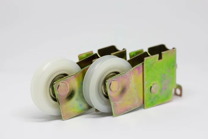 Two white plastic wheels with metal brackets are positioned side by side on a plain background. The yellowish, slightly iridescent finish on the metal parts hints at their function—likely key components in the repair of a sliding glass door mechanism.