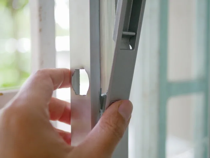 A hand adjusts a metal window frame, with part of the frame slightly detached to reveal a locking mechanism—similar to those used in sliding doors—against a blurred background. This common scene could be found in homes across Tampa, FL.