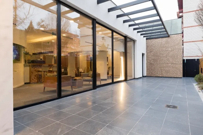 Spacious modern patio with sleek black tiled floor and large glass sliding doors revealing a cozy interior with armchairs and decor. A pergola with beams extends overhead, and a textured wall is visible in the background, creating an inviting space typical of stylish Tampa, FL homes.