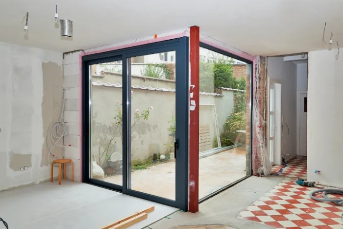 A room under renovation in Tampa, FL, features large sliding glass doors opening to a small outdoor garden. The room has unfinished walls, exposed wires, and a red and white checkered floor. A wooden stool and construction materials are scattered around as local experts work on the repair.