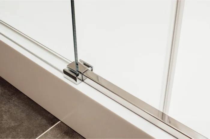 Close-up of a modern glass sliding door with a chrome hinge mounted on a white frame. The flooring consists of dark tiles, and the image focuses on the minimalistic design and sleek lines crafted by a local company.