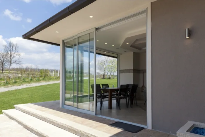 Sliding glass doors open to reveal a dining area inside a modern house in Tampa, FL. Outside, a patio overlooks a grassy lawn and trees under a partly cloudy sky. Steps lead up to the entrance, creating a seamless indoor-outdoor transition in this charming local setting.