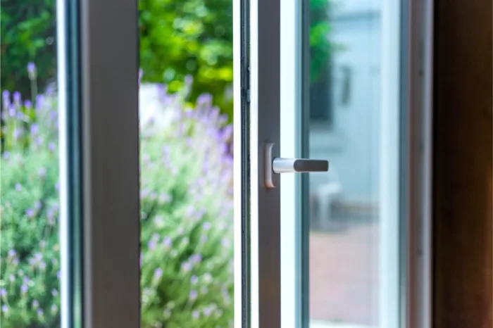 An open sliding glass door leads to a garden with blooming lavender and greenery, allowing sunlight to brighten the indoor space. The metallic handle is visible, indicating the door is slightly ajar and in need of repair.