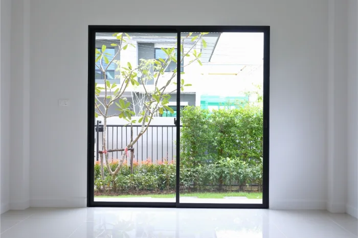 A large sliding glass door opens to a view of a small garden with a tree and neatly trimmed shrubs. Beyond the garden, there is a black metal fence and a modern building. This serene retreat in Tampa, FL, features an interior with a glossy white floor that reflects the abundant natural light.