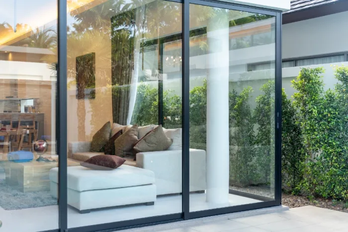 Luxurious living room viewed through large sliding glass doors, featuring a white sectional sofa with brown and beige cushions. Lush green shrubs and trees are visible outside, reflecting on the pristine glass in this stunning Tampa, FL home.