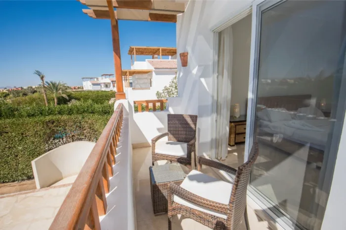 The sunlit balcony, featuring wicker chairs and a small table, offers a serene garden view. White walls and wooden railings complement the setting. A sliding glass door seamlessly connects to the bedroom, where local scenery of clear blue skies and distant trees enhances the tranquil ambiance.