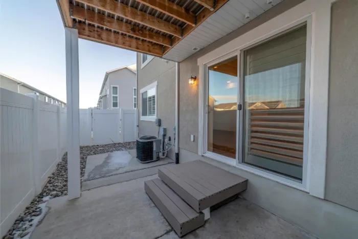 View of a small backyard patio in Tampa, FL, featuring a wooden deck and sliding glass door leading into the house. The area is enclosed by a white fence, with an air conditioning unit beside the concrete path. Some rocks accent the landscaping, offering a tranquil oasis.