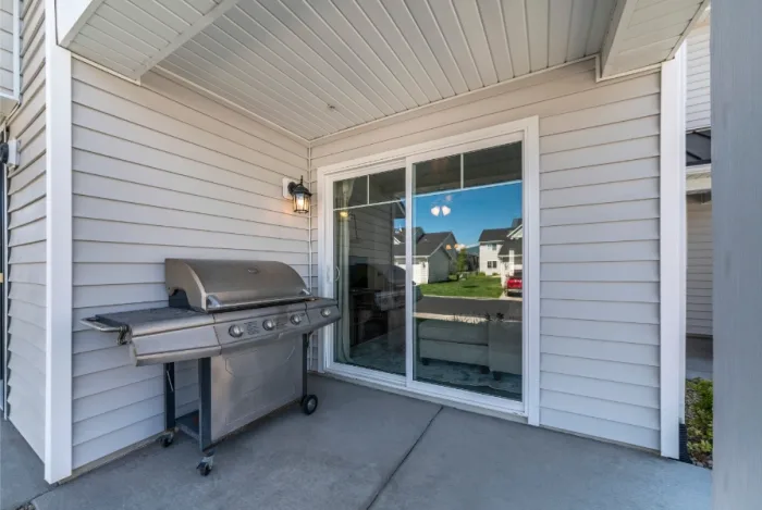 A covered patio area with a stainless steel grill on the left and a sliding glass door from a local company on the right. The patio is surrounded by white siding, with neighboring houses visible in the background through the glass door.