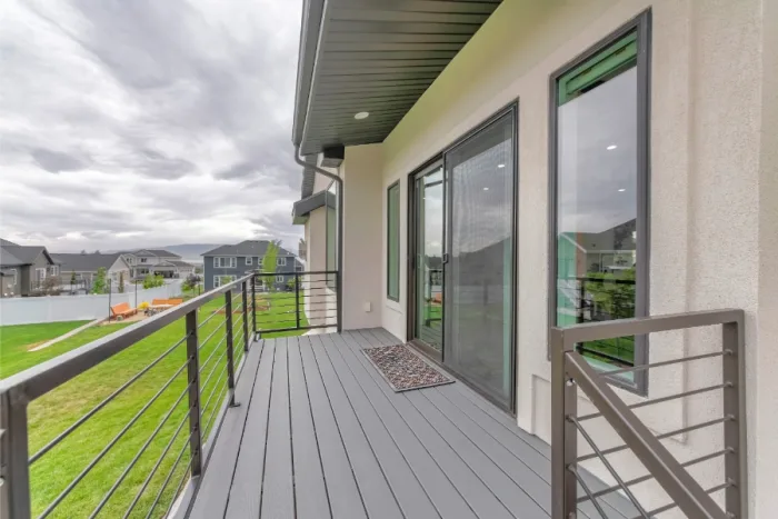 A contemporary residential balcony with gray decking and black railing overlooks a grassy lawn and neighboring houses. Large sliding glass doors provide access to the modern interior, where a textured doormat lies near the entrance. A cloudy sky looms in the background, casting a serene ambiance.