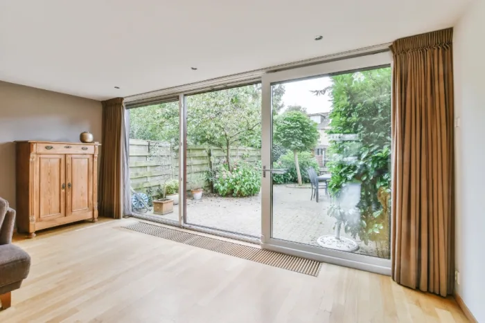 A spacious living room with wooden flooring features a sliding glass door opening to a garden patio. The room, located in Tampa, FL, has brown curtains and a wooden cabinet on the left. The patio area is surrounded by greenery, with outdoor seating visible for local gatherings.