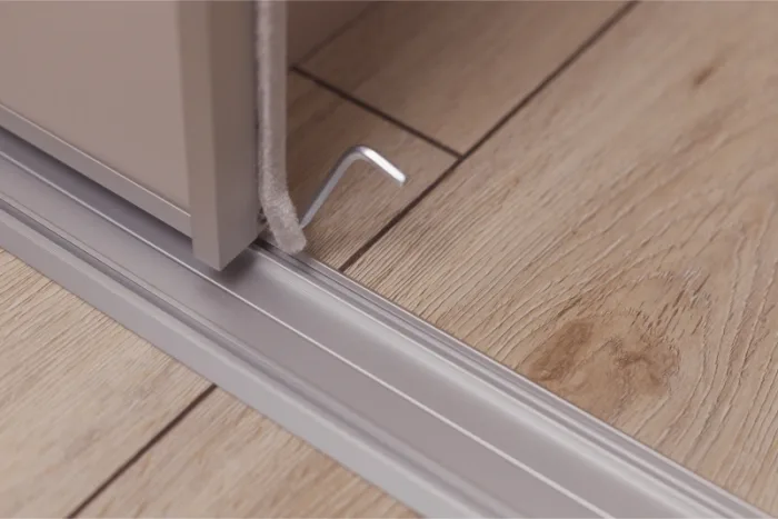 Close-up of a sliding glass door track on a wooden floor. A small metal hook protrudes from the base of the door, perfectly positioned above the track. The scene captures the intersection of the sliding glass door, track, and flooring with precision.