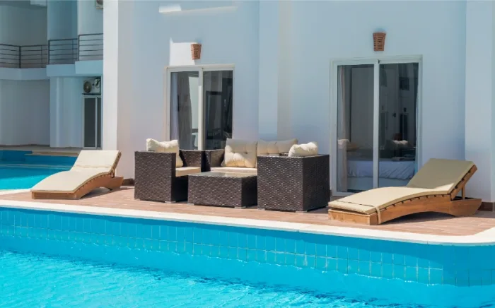 A modern outdoor patio in Tampa, FL features a dark wicker sofa set and two cushioned lounge chairs beside a blue swimming pool. The white building in the background, representative of a local architectural style, boasts large glass doors and windows.