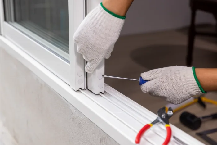 A person wearing white gloves uses a screwdriver to adjust a sliding window, possibly in preparation for a local repair task. A pair of pliers rests on the window sill, hinting at an ongoing setup. The scene unfolds indoors with muted lighting, creating a focused atmosphere.