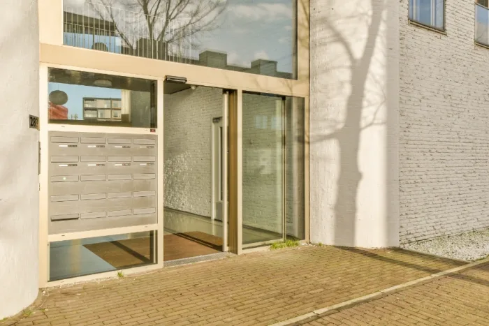 A modern building entrance with large sliding glass doors and a set of mailboxes mounted on the white brick wall. The shadow of a tree is cast on the wall, and the sun shines on the scene. The pavement is made of bricks, reflecting a sleek company aesthetic.