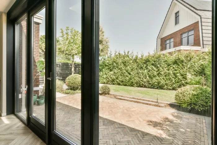 View of a sunny backyard through large glass doors, featuring a paved patio, neatly trimmed grass, and a tall hedge. A modern two-story house is visible in the background, bordered by trees and plants—a perfect setting for local gatherings or repair projects in Tampa FL.