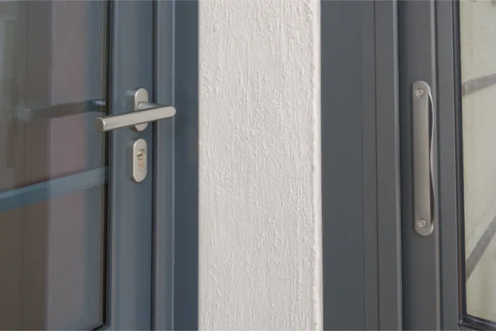 Close-up of a gray door handle and lock on a sliding glass door next to a long vertical handle on a white wall panel. Reflections shimmer on the glass, highlighting modern design elements.