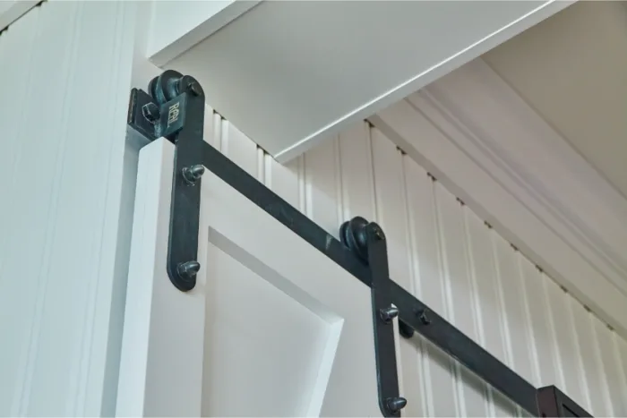 Close-up of a white barn door with a black sliding rail system, reminiscent of a sliding glass door, mounted on a white beadboard wall. The hardware includes visible rollers and brackets, accentuating the modern rustic aesthetic of the doorway.